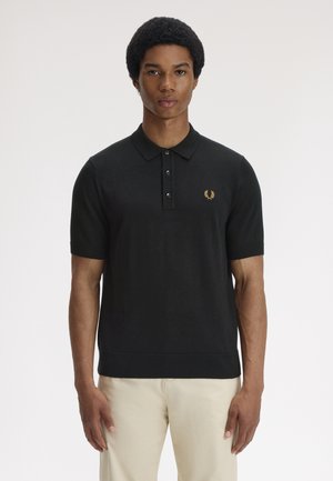Jeune homme aux cheveux courts bouclés portant un polo noir à manches courtes avec un logo brodé et un pantalon beige clair, debout immobile.