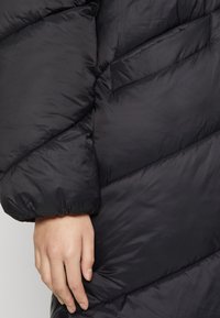 Svart puffjacka med diagonalt quiltad sömnad, mjuk textur och en något rynkad ärmslut. Hand som vilar på tyget.