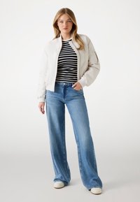 Giacca bomber beige chiaro con polsini a costine, indossata sopra una camicia a righe blu e bianche. Abbinata a jeans blu a gamba larga e sneakers bianche.