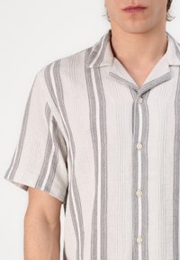 Camisa de manga corta abotonada en tejido gris claro texturizado con rayas verticales en gris oscuro y blanco, y un cuello con muesca.