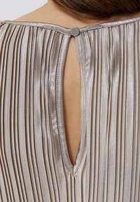 Tejido plisado de color beige claro con un cierre trasero en forma de ojal, que presenta un pequeño detalle de botón. Textura suave y acabado brillante.