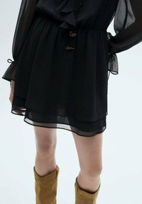 Black sheer dress featuring a ruffled neckline, waist tie, and layered hem. Brown knee-high boots are visible below.