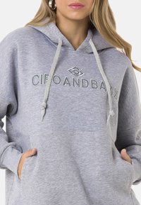 Grauer Kapuzenpullover mit großer Vorderentasche und dekorativem Text. Weicher Stoff, mit Kordeln zum Zuziehen und einem locker sitzenden Schnitt.