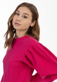 Top fucsia a maniche lunghe in tessuto morbido, con scollo tondo e maniche voluminose, indossato da un modello su uno sfondo bianco.