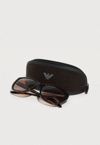 Γυαλιά ηλίου με σκελετό από καβούκι χελώνας και σκουρόχρωμους φακούς, τοποθετημένα δίπλα σε μαύρη σουέντ θήκη με φερμουάρ που φέρει ασημένια λεπτομέρεια λογότυπου.
