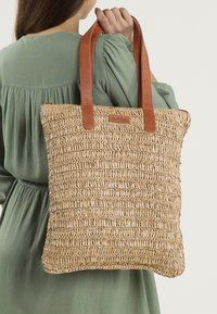 Geweven rieten tote bag met een rechthoekige vorm, tan leren handvatten en een gestructureerd oppervlak. Bevat een gelabelde leren tag aan de voorkant.