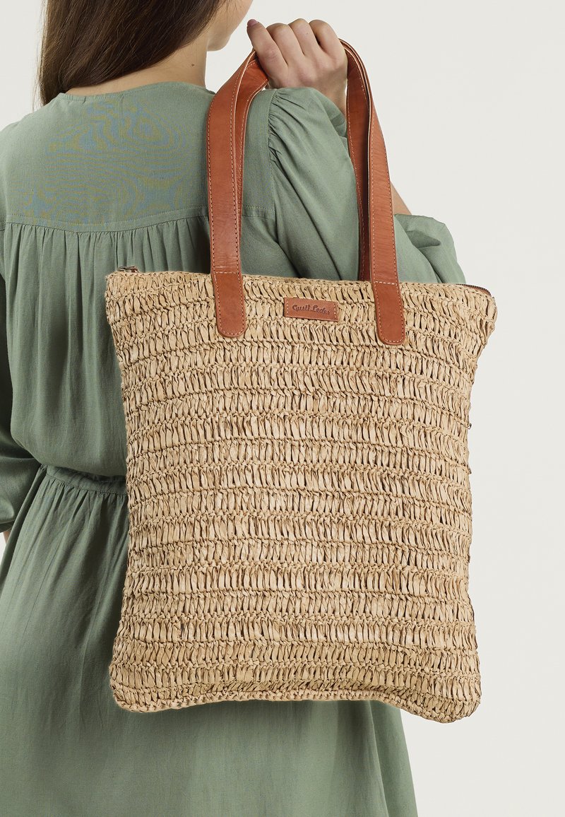 Geweven rieten tote bag met een rechthoekige vorm, tan leren handvatten en een gestructureerd oppervlak. Bevat een gelabelde leren tag aan de voorkant.