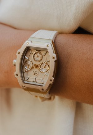 Montre en silicone blanche avec un boîtier en métal doré, en forme octogonale, trois petits cadrans et des détails en or texturé sur le cadran.
