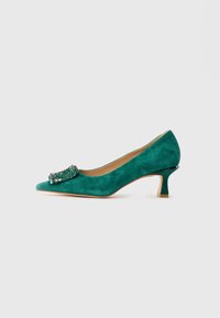 Scarpa verde in suede con tacco kitten e punta affusolata, decorata con una fibbia gioiello sul davanti, mostrata in profilo laterale su sfondo bianco.