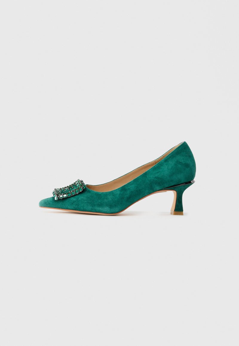 Scarpa verde in suede con tacco kitten e punta affusolata, decorata con una fibbia gioiello sul davanti, mostrata in profilo laterale su sfondo bianco.