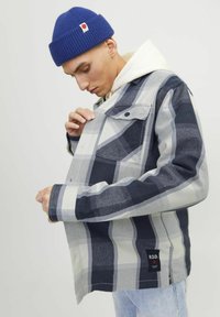 Giacca a quadri nei toni del blu navy, grigio e bianco, realizzata in tessuto intrecciato con tasca sul petto e etichetta R.D.D. Indossata sopra un hoodie color crema.