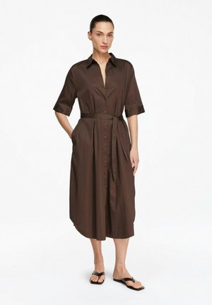 Kobieta z włosami związanymi do tyłu nosi brązową, zapinaną na guziki sukienkę midi z paskiem, z rękawami do łokci, oraz czarne sandały z odkrytymi palcami, stoi z rękami w kieszeniach.