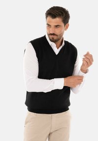 Gilet nero in maglia con scollatura a V, indossato sopra una camicia bianca a maniche lunghe. Presenta un orlo e delle aperture per le braccia a coste, e una texture liscia.