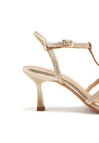 Sandaia dorada con tiras, textura trenzada, punta abierta y tacón delgado. Presenta un acabado metálico y una pequeña hebilla ajustable.