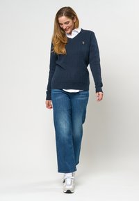 Navy kabelgebreide trui met een V-hals en geribde manchetten, gecombineerd met blauwe denim jeans met een relaxte pasvorm en witte sneakers.