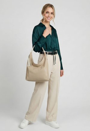 Jonge vrouw glimlacht, draagt een donkergroene blouse, beige broek, witte sneakers, houdt een beige schoudertas met kwastje vast tegen een neutrale achtergrond.