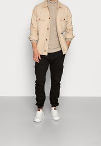 Mann in beigem Jackett, beige Strickrollkragenpullover, schwarzen Cargo-Hosen und weißen Sneakers, der vor einer schlichten Wand steht.
