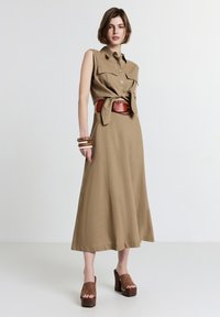 Abito khaki senza maniche con bottoni frontali, vita cintata e tasche laterali, abbinato a sandali con piattaforma marroni e bracciali di legno.