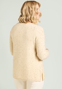 Beige trui met een zachte textuur, versierd met verspreide metalen accenten, voorzien van een ronde hals en zijslitten voor bewegingsvrijheid.