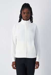 Felpa bianca con zip e colletto alto, maniche raglan. Tessuto morbido, dettaglio logo discreto sul petto, abbinata a pantaloni scuri.