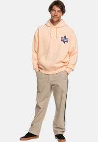 Sudadera melocotón con un bolsillo frontal y un logo azul, combinada con pantalones de pana beige que tienen una franja negra. Zapatillas negras completan el conjunto.