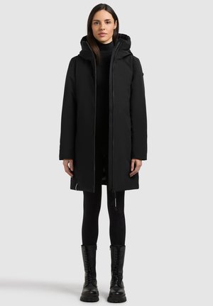 Femme portant un long manteau matelassé noir avec capuche, des leggings noirs et des bottes noires à lacets, debout devant un fond clair uni.