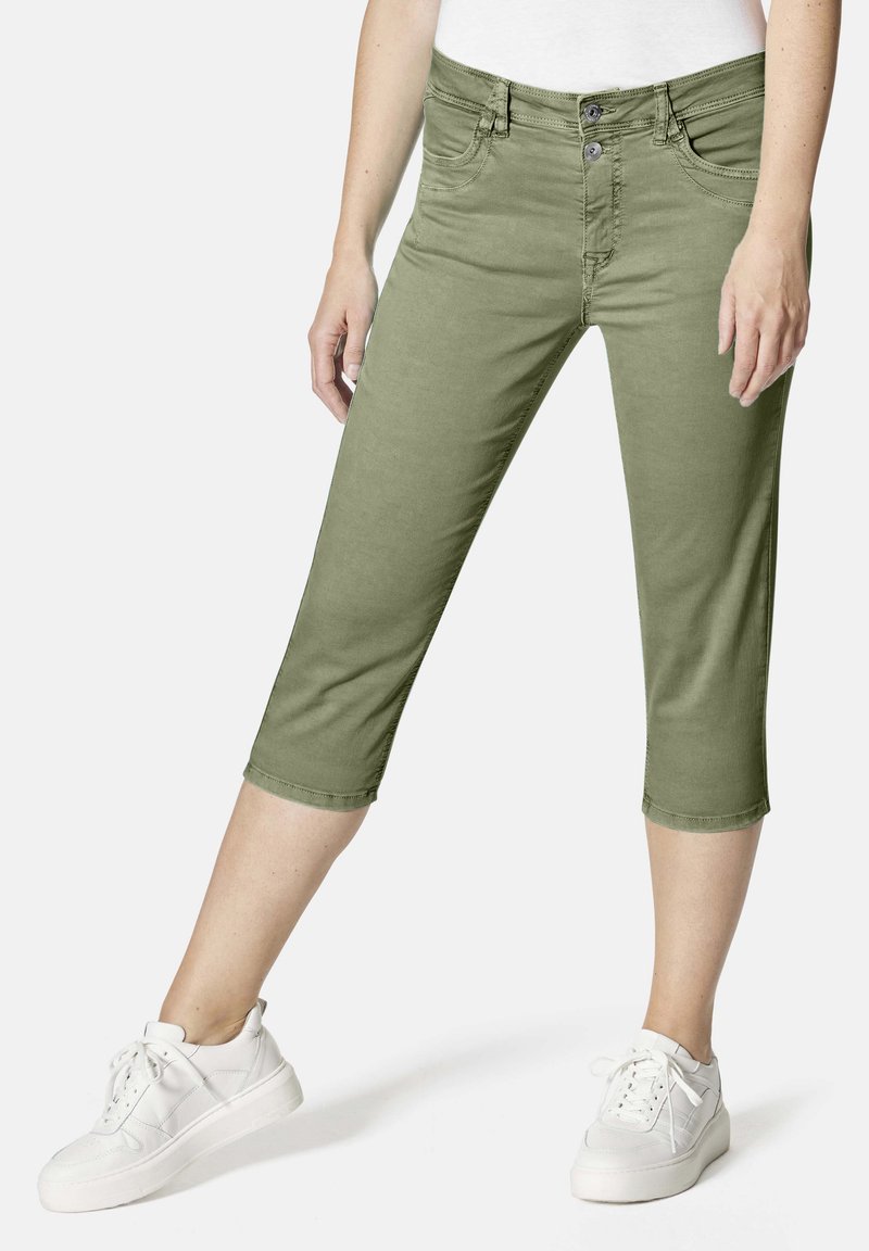 Olivgrüne, cropped Hose mit frontalen Taschen, Reißverschluss und Zwei-Knopf-Verschluss; kombiniert mit weißen Sneakers. Glattes Baumwollgemisch.