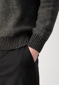 Personne portant un pull en maille gris foncé avec des poignets côtelés, main partiellement dans la poche d'un pantalon noir sur un fond neutre.