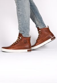 Zapatillas altas de piel marrón con suelas de goma blanca, cuentan con costuras de acento y ojales de metal. Piel texturizada con detalle del logo en el costado.