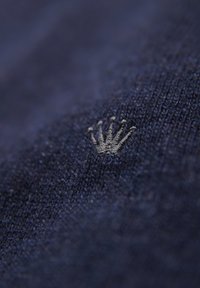 Marinefarbene Strickware mit einer strukturierten Oberfläche, die in der unteren rechten Ecke ein kleines, silbernes Kronenlogo aufgestickt hat.