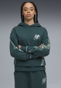 Sudadera verde oscuro con un logo delantero y patrones blancos intrincados en las mangas. Presenta un corte relajado y una textura suave.