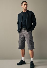 Männeroutfit mit einer schwarzen Bomberjacke, einem marineblauen T-Shirt, grauen Cargo-Shorts mit Taschen und schwarzen Turnschuhen mit weißen Socken.