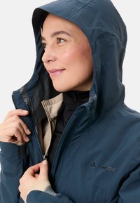 Marineblauwe waterdichte jas met een capuchon, volledige rits en binnenvoering met een geometrisch patroon. Verstelbare manchetten en logo op de borst.