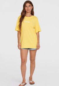 Geel katoenen T-shirt met korte mouwen, met de witte tekst "The Future Surf Society" op de voorkant. Gecombineerd met denimshorts.