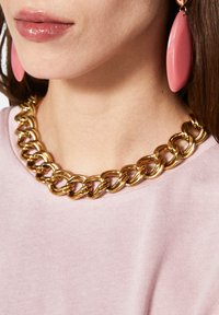Collana di catena d'oro con grandi anelli indossata sopra una blusa rosa chiaro, completata da orecchini a goccia rosa.