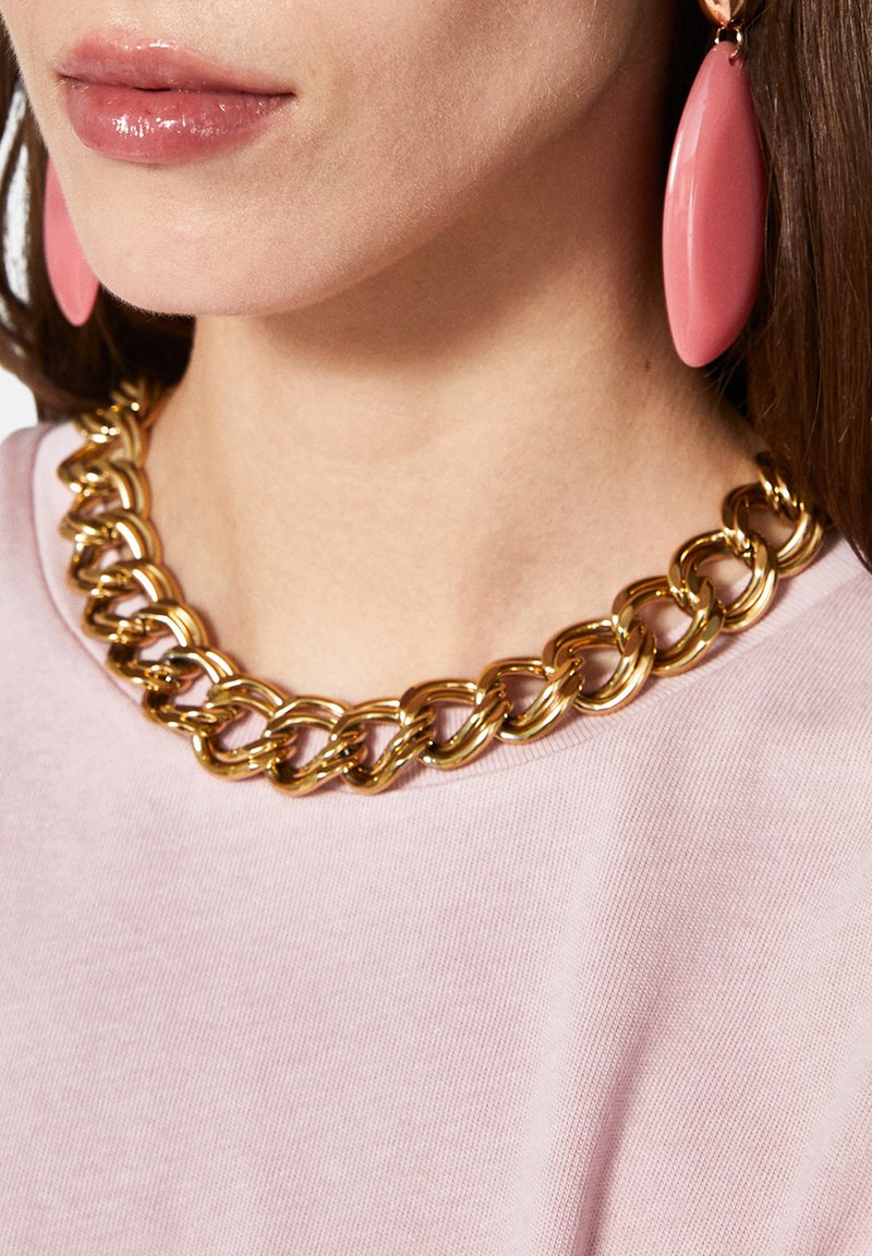 Collana di catena d'oro con grandi anelli indossata sopra una blusa rosa chiaro, completata da orecchini a goccia rosa.