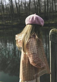 Półprzezroczysta różowa kurtka w kratkę z falbaniastymi rękawami, w komplecie z różowym wełnianym beretem, noszona przez postać stojącą w pobliżu zbiornika wodnego.