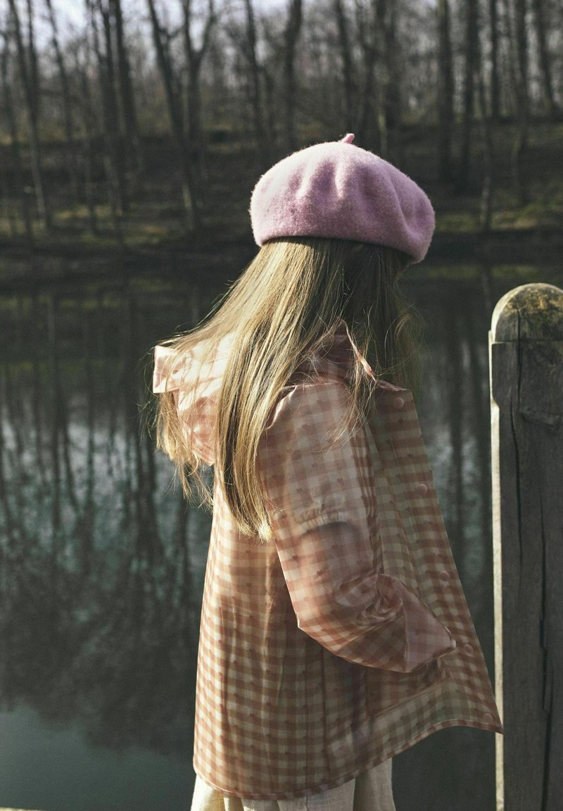 Półprzezroczysta różowa kurtka w kratkę z falbaniastymi rękawami, w komplecie z różowym wełnianym beretem, noszona przez postać stojącą w pobliżu zbiornika wodnego.
