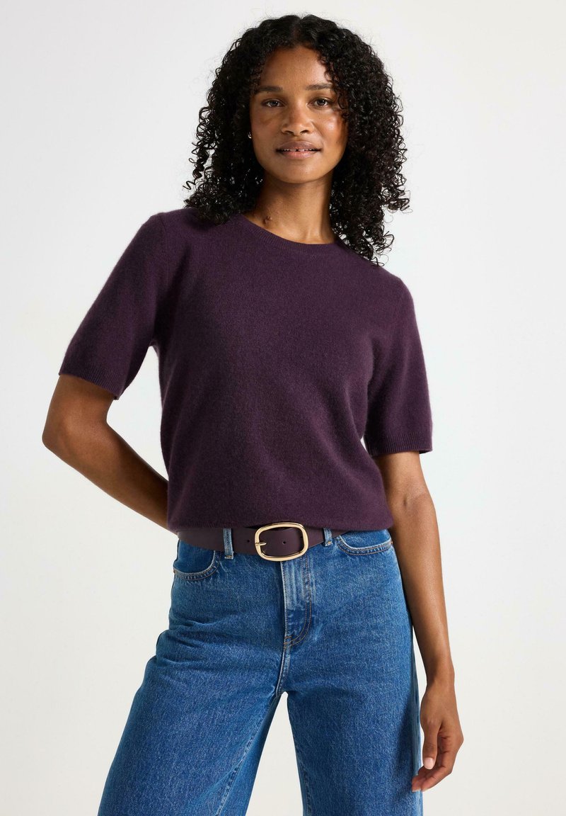 Maglione viola a maniche corte realizzato in materiale morbido, abbinato a jeans a vita alta blu chiaro e una cintura marrone con fibbia dorata.