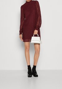 Vestido de encaje burdeos con mangas largas y abullonadas y un cuello alto. Combinado con un pequeño bolso blanco y zapatos negros con calcetines.