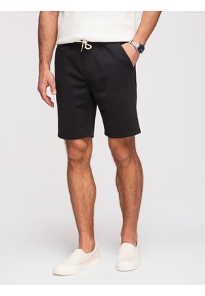 Mężczyzna w czarnych szortach ze sznurkiem, białych butach wsuwanych i białej, bezrękawnikowej koszulce, stojący z jedną ręką w kieszeni.