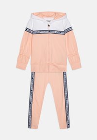 Barack és fehér sportöltöny cipzáros kapucnis pulóverrel, amelyen sötétkék logós szalag található. Tartalmazza a hozzá illő leggingset oldalsó logó részletekkel és rugalmas mandzsettákkal.