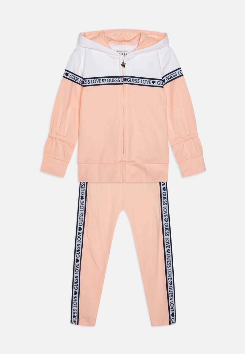 Barack és fehér sportöltöny cipzáros kapucnis pulóverrel, amelyen sötétkék logós szalag található. Tartalmazza a hozzá illő leggingset oldalsó logó részletekkel és rugalmas mandzsettákkal.