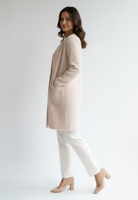 Beżowy płaszcz z mieszanki wełny o prostym kroju, z dwoma przednimi kieszeniami i fakturowaną powierzchnią, w zestawieniu z białymi spodniami i beżowymi butami na klocku.