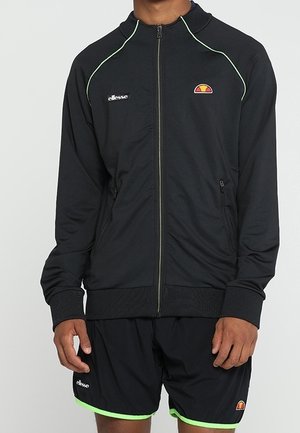 Mann trägt schwarze Sportjacke mit Reißverschluss und passende Shorts mit limonengrünem Rand, beide mit Ellesse-Logos.