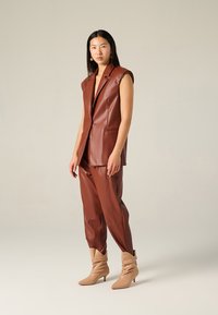 Blazer senza maniche in finta pelle marrone e pantaloni coordinati, con un design strutturato. Stivaletti beige morbidi alla caviglia con punta affilata.