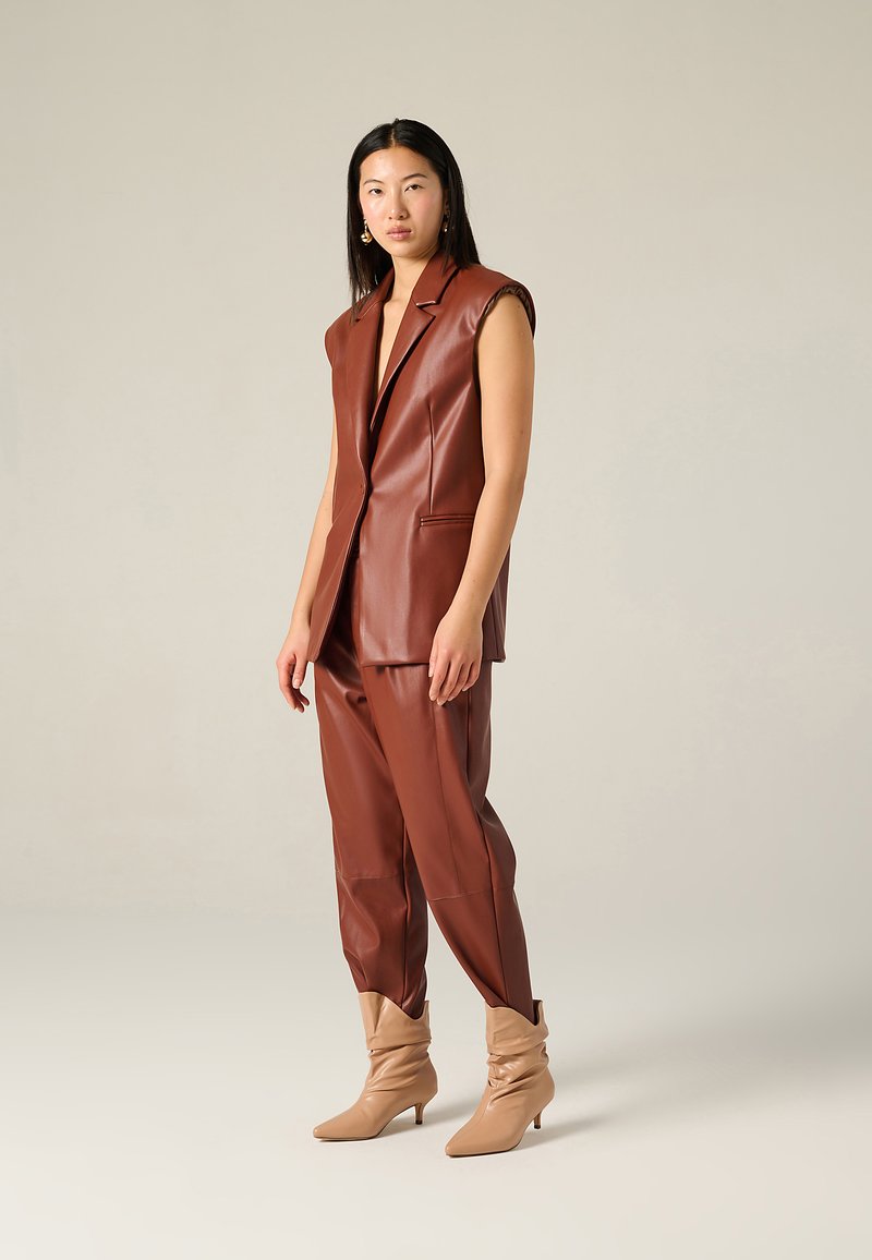 Blazer senza maniche in finta pelle marrone e pantaloni coordinati, con un design strutturato. Stivaletti beige morbidi alla caviglia con punta affilata.