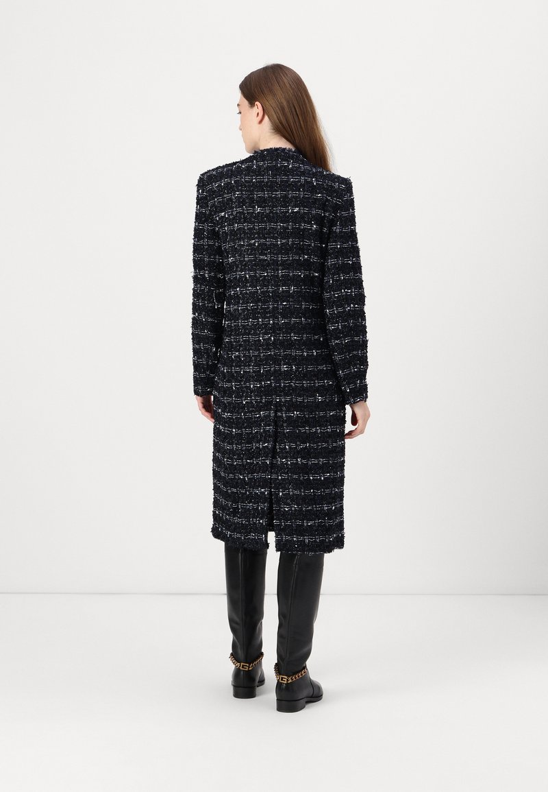 Langer, schwarzer Tweed-Mantel mit strukturiertem Muster, geradem Schnitt und leicht fransigen Kanten, getragen über schwarzen knielangen Stiefeln.