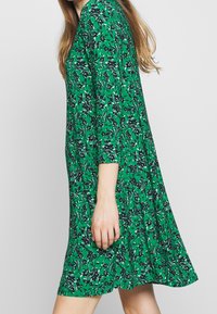 Green floral dress with three-quarter sleeves, featuring a flowing design. Material appears lightweight with a vivid floral pattern in navy and white.