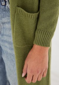 Olive-zielony dziany kardigan z prążkowanymi mankietami, miękką teksturą i boczną kieszenią. W tle widać denimu.